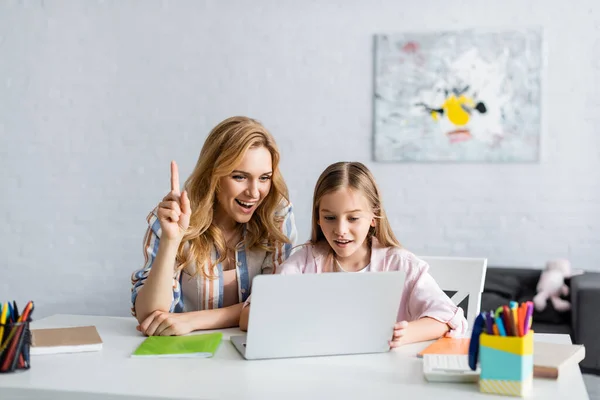 Selective focus of smiling woman having idea while using laptop near kid and stationery on table — Stock Photo