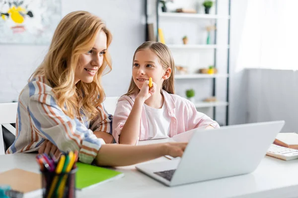 Selective focus of smiling woman using laptop near kid holding pen during online education — Stock Photo