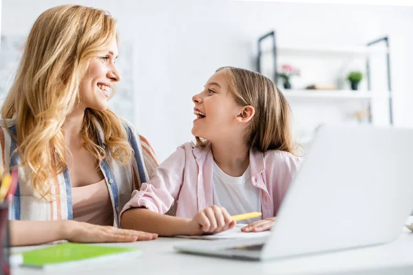 Concentration sélective de la mère souriant à sa fille près de la papeterie et de l'ordinateur portable sur la table à la maison — Photo de stock