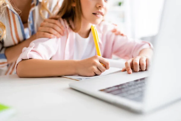 Ausgeschnittene Ansicht einer Frau, die ein Kind umarmt, das auf einem Notizbuch neben dem Laptop auf dem Tisch schreibt — Stockfoto