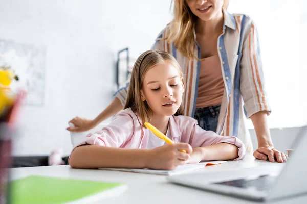 Selective focus of smiling mother standing near daughter writing on notebook near laptop on table — Stock Photo