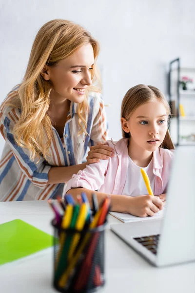 Selektiver Fokus des Kindes, das während des elektronischen Lernens in der Nähe einer lächelnden Mutter auf einem Notizbuch schreibt — Stockfoto