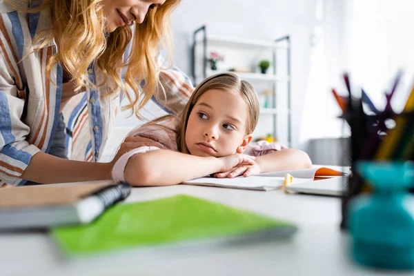 Selective focus of mother embracing sad daughter near stationery on table — Stock Photo