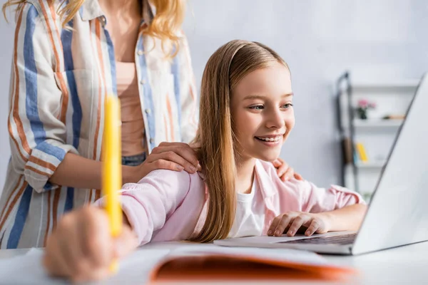 Enfoque selectivo de la mujer abrazando sonriente hija escribiendo en el portátil cerca de la computadora portátil en casa - foto de stock