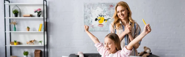 Panoramic shot of smiling mother standing near cheerful kid holding pen at home — Stock Photo