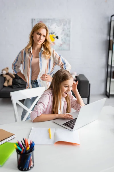 Selektiver Fokus wütender Mutter, die neben aufgeregtem Kind steht und Laptop in der Nähe von Schreibwaren auf dem Tisch benutzt — Stockfoto