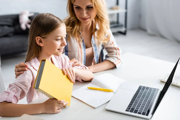 Selektiver Fokus der Frau, die lächelnde Tochter umarmt, die Buch neben Laptop und Notizbuch auf dem Tisch hält — Stockfoto