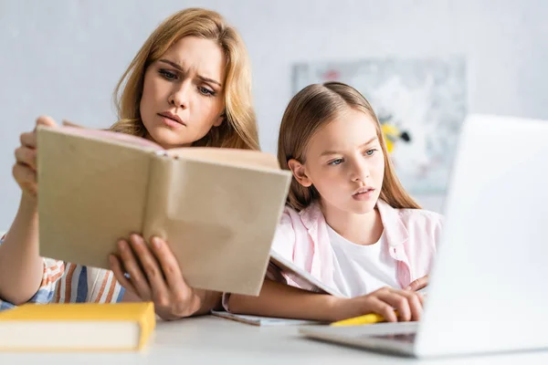 Вибіркове зосередження уваги зосередженої жінки на читанні книжки з дитиною за допомогою ноутбука за столом. — стокове фото