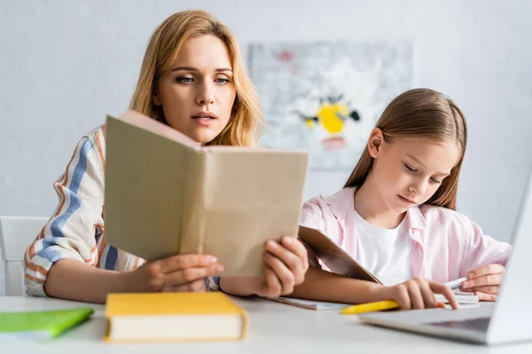 Concentration sélective de livre de lecture de femme tout en aidant la fille pendant l'éducation en ligne à la maison — Stock Photo
