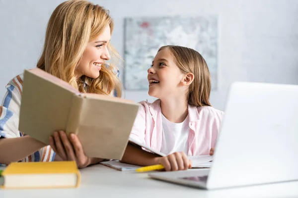 Selective focus of cheerful child looking at mother with book near laptop at table — Stock Photo