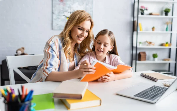 Selektiver Fokus einer lächelnden Frau, die ein Notizbuch in der Hand hält, während sie ihrer Tochter bei der Online-Ausbildung zu Hause hilft — Stockfoto