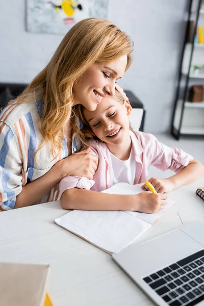 Селективный фокус женщины обнимающей улыбающуюся дочь возле канцелярских принадлежностей и ноутбука на столе дома — стоковое фото