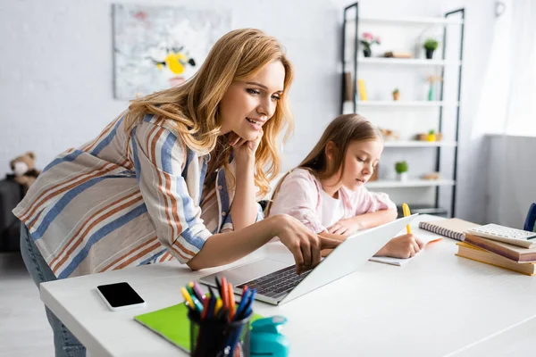 Enfoque selectivo de la madre sonriente apuntando con el dedo a la computadora portátil cerca del niño escribiendo en el cuaderno en casa - foto de stock