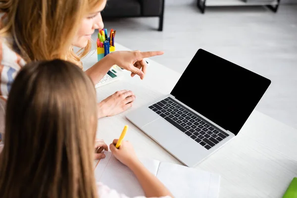 Selective focus of woman pointing with finger at laptop near kid writing on notebook during electronic learning at home — Stock Photo