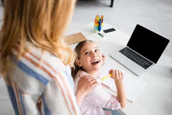 Selektiver Fokus des positiven Kindes, das Mutter ansieht, während es Stift in der Nähe von Notizbuch und Laptop auf dem Tisch hält — Stockfoto