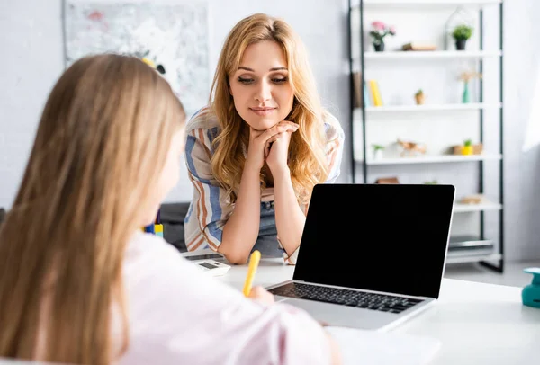 Selective focus of mother looking at daughter writing on notebook near laptop on table — Stock Photo