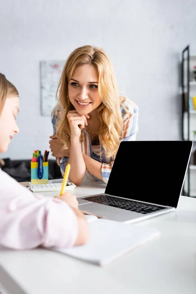 Selective focus of smiling woman looking at kid writing on copybook near laptop with blank screen — Stock Photo