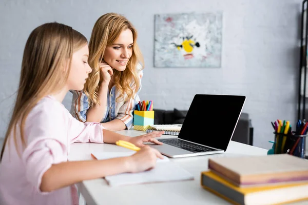 Selective focus of smiling woman standing near kid using laptop during electronic learning at home — Stock Photo