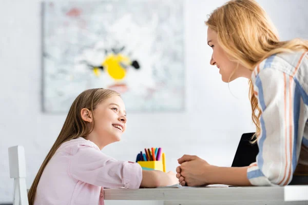 Vista lateral del niño sonriente mirando a la madre cerca de la computadora portátil y papelería en la mesa — Stock Photo