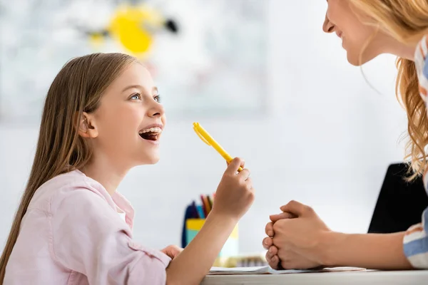 Вид сбоку позитивного ребенка, держащего ручку и смотрящего на мать возле тетради на столе — стоковое фото