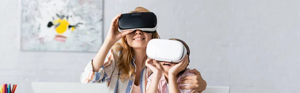 Panoramic shot of smiling mother and daughter using vr headsets near laptop at home — Stock Photo