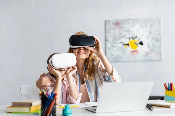 Enfoque selectivo de la mujer sonriente abrazando a la hija mientras usa auriculares de realidad virtual cerca de computadoras portátiles y libros - foto de stock