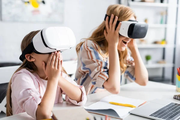 Selektiver Fokus aufgeregter Kinder, die Mund bedecken, während sie VR-Headsets mit Mutter in der Nähe von Laptop und Schreibwaren verwenden — Stockfoto