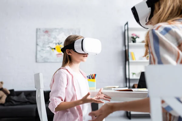 Foco seletivo da criança usando fones de ouvido vr perto da mãe e papelaria na mesa — Fotografia de Stock