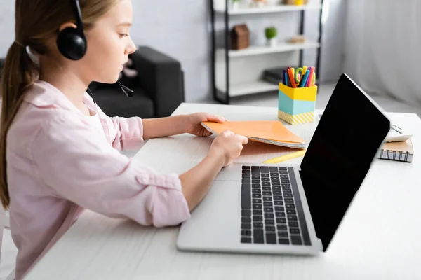 Side view of kid in headset taking notebook near laptop on table — Stock Photo