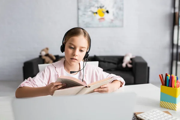 Selective focus of child in headset reading book near laptop on table — Stock Photo