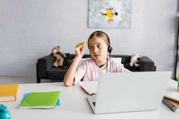 Selective focus of pensive child holding pen while using headset near laptop and stationery on table — Stock Photo