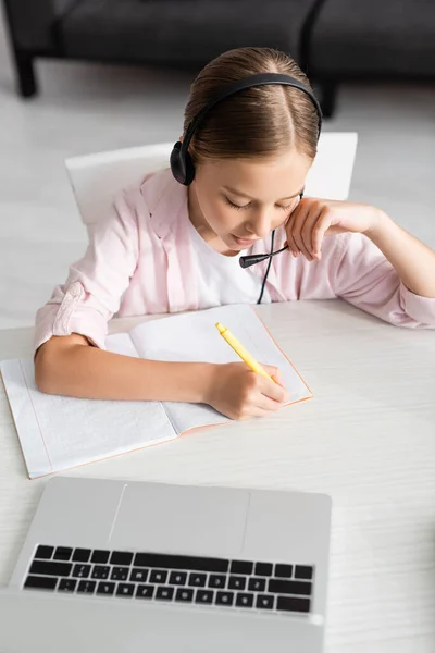 Вид сверху на ребенка в гарнитуре записи на ноутбуке рядом с ноутбуком на столе — стоковое фото