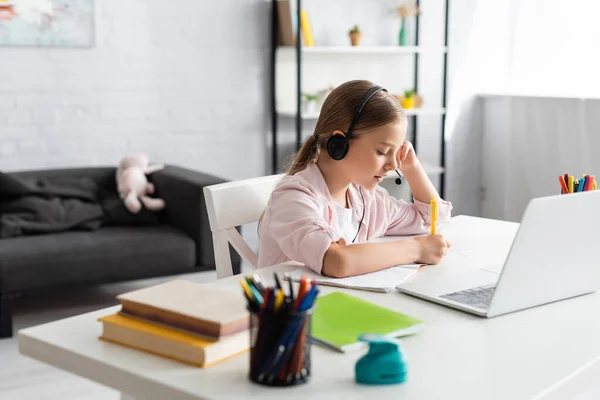 Selektiver Fokus des Kindes beim Schreiben auf dem Notizbuch und beim Benutzen eines Headsets in der Nähe des Laptops auf dem Tisch — Stockfoto