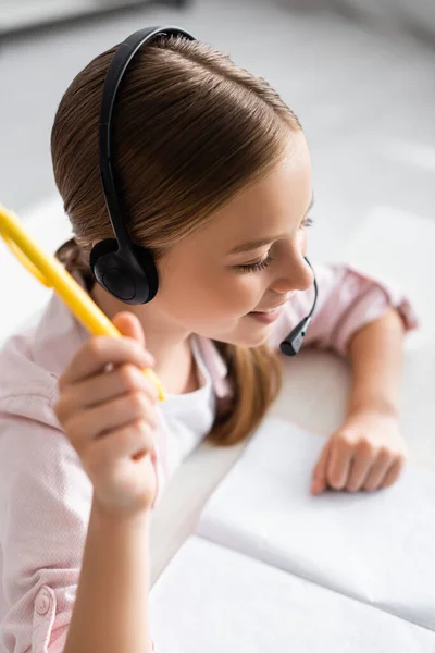 Blick von oben auf lächelndes Kind im Headset, das Stift neben Notizbuch auf dem Tisch hält — Stockfoto