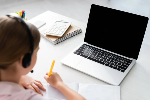 Selective focus of child in headset writing on notebook near laptop and stationery on table — Stock Photo
