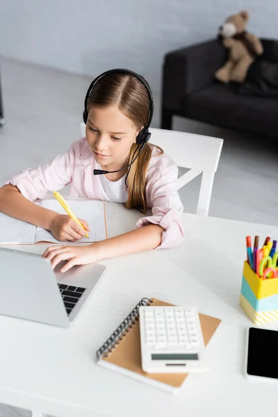 Selektiver Fokus des Kindes beim Schreiben auf dem Notizbuch, während es Headset und Laptop auf dem Tisch benutzt — Stockfoto