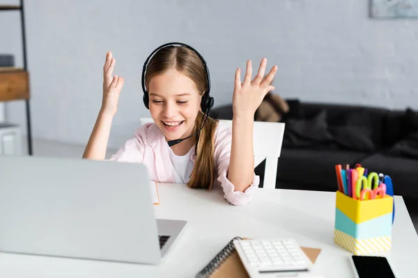 Селективный фокус позитивного ребенка в наушниках, смотрящего на ноутбук рядом со стационарным столом — стоковое фото
