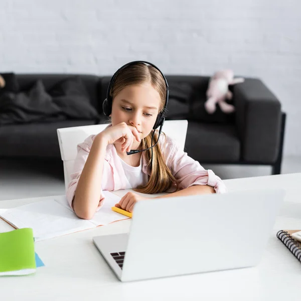 Вибірковий фокус милої дитини в гарнітурі, що сидить поруч з ноутбуком і ноутбуком на столі — стокове фото