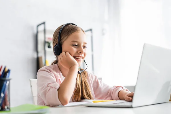 Selective focus of smiling kid using headset and laptop at table — Stock Photo