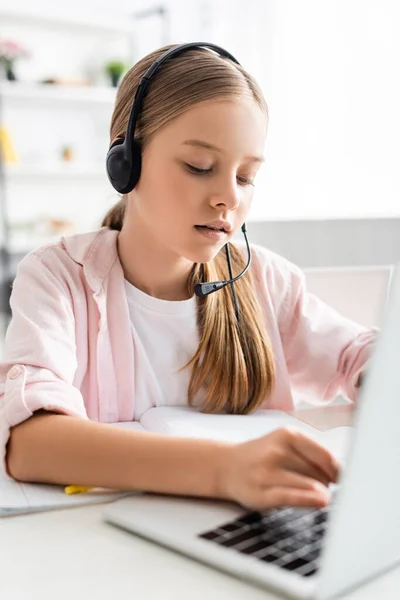 Selective focus of cute child in headset typing on laptop near notebook on table — Stock Photo