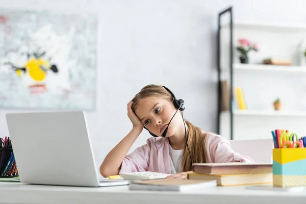 Selective focus of tired kid looking at laptop while using headset near books on table — Stock Photo