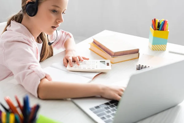 Enfoque selectivo del niño con auriculares usando computadora portátil y calculadora durante la educación en línea - foto de stock