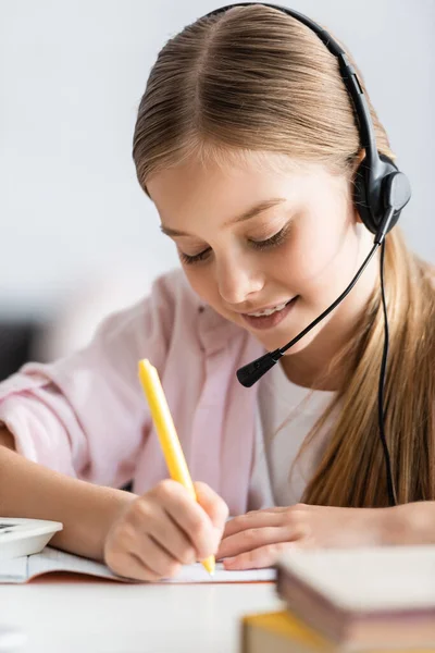 Selective focus of smiling child in headset writing on copy book at table — Stock Photo