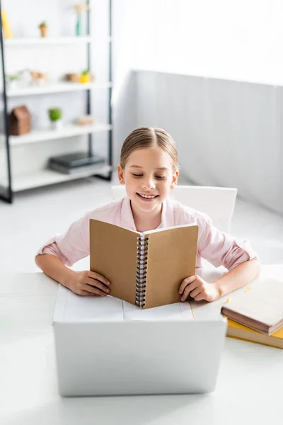 Enfoque selectivo del niño sonriente sosteniendo un cuaderno cerca de la computadora portátil y libros en la mesa - foto de stock