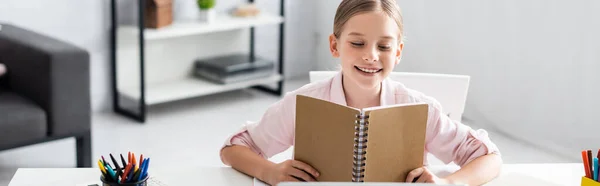 Prise de vue panoramique d'un enfant souriant tenant un carnet près de la papeterie à table — Photo de stock