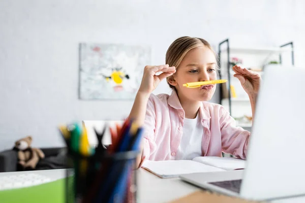 Selective focus of kid holding pen near mouth during webinar on laptop at home — Stock Photo