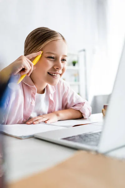 Выборочный фокус улыбающегося ребенка, держащего ручку и смотрящего на ноутбук на столе — стоковое фото