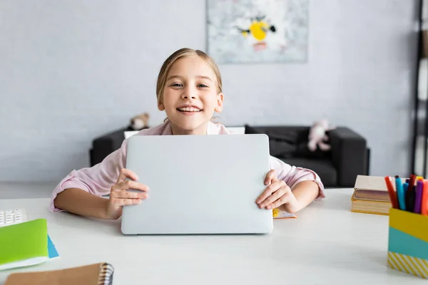 Enfoque selectivo del niño sonriente sosteniendo la computadora portátil y mirando a la cámara cerca de papelería - foto de stock