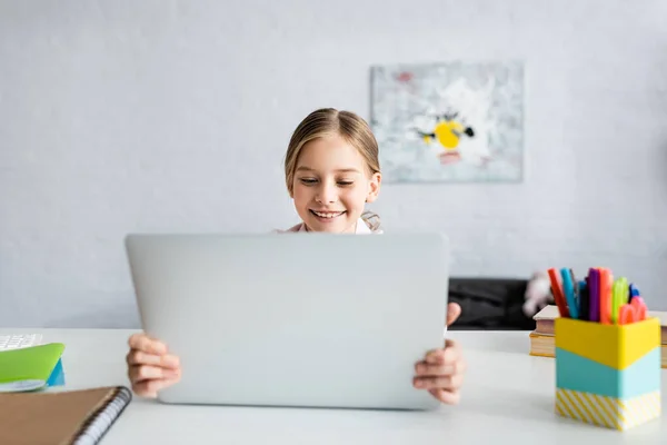 Enfoque selectivo del niño sonriente sosteniendo la computadora portátil cerca de libros en la mesa - foto de stock