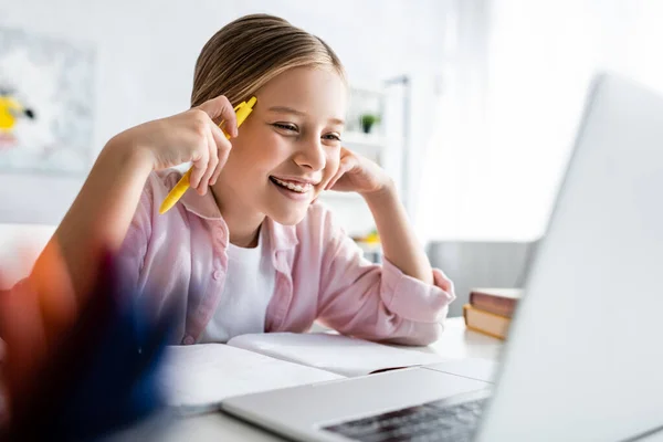 Focus selettivo del bambino positivo che tiene la penna vicino al notebook e guarda il computer portatile sul tavolo — Foto stock